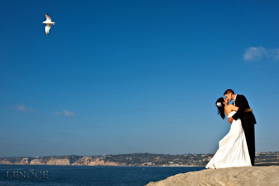 Bride and groom embracing on a cliff against a clear blue sky.